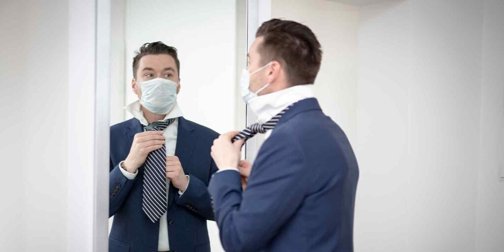 man in a medical mask corrects his tie in front of the mirror during the covid-19 pandemic