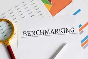 text BENCHMARKING is written on a paper with a pen and a magnifying glass on the table